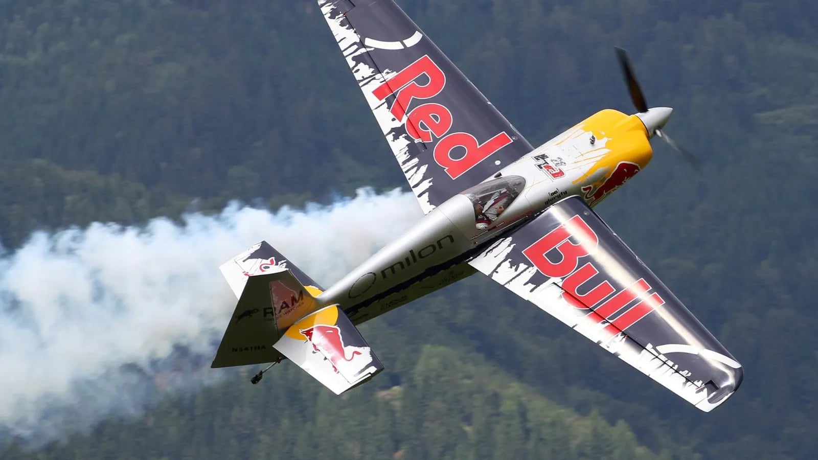 A Red Bull branded race plane trailing smoke over a green valley.
